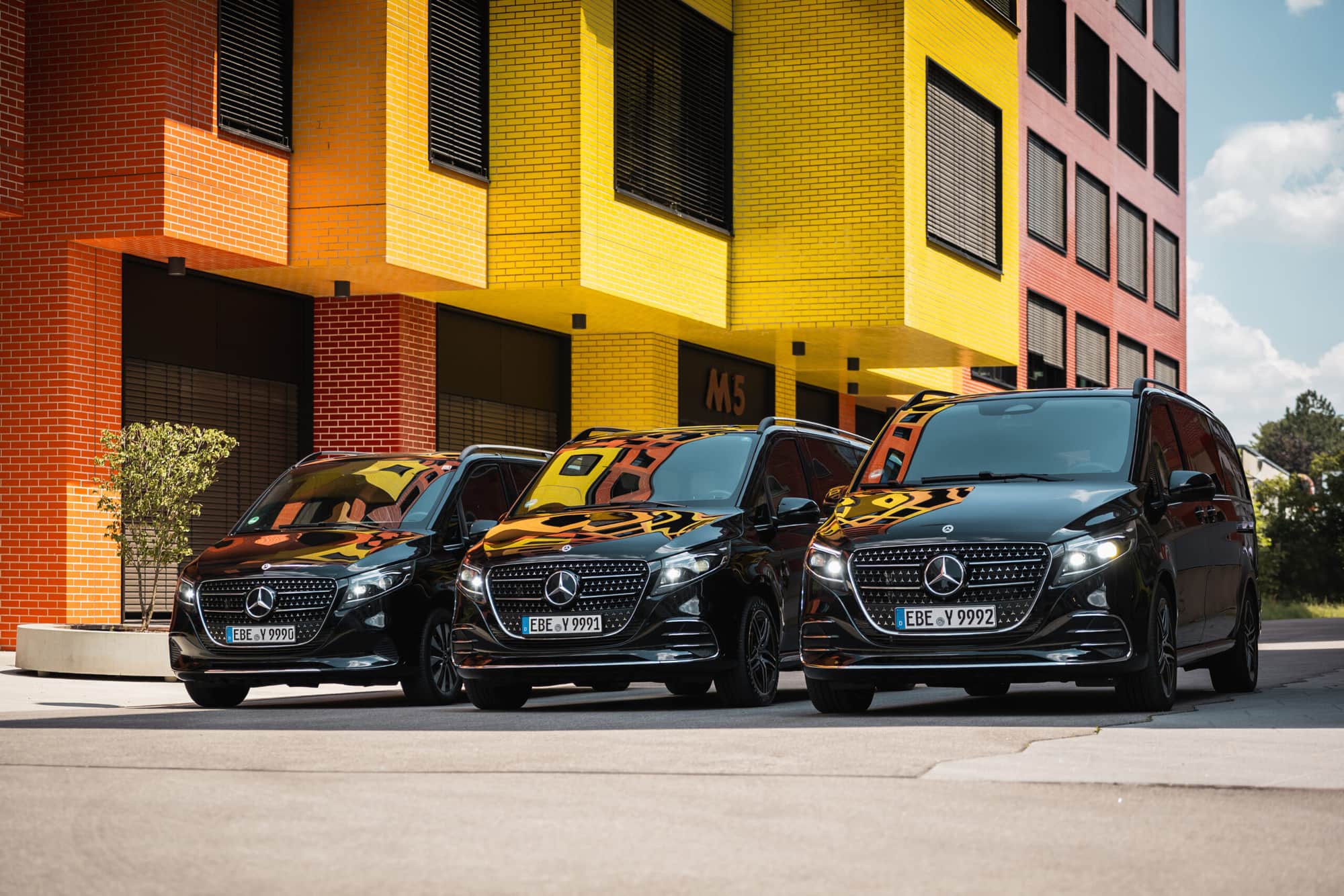Three black AYDEN Mercedes-Benz V-Class vans outside an office building with a colourful brick façade – vehicles for the Allianz Arena shuttle