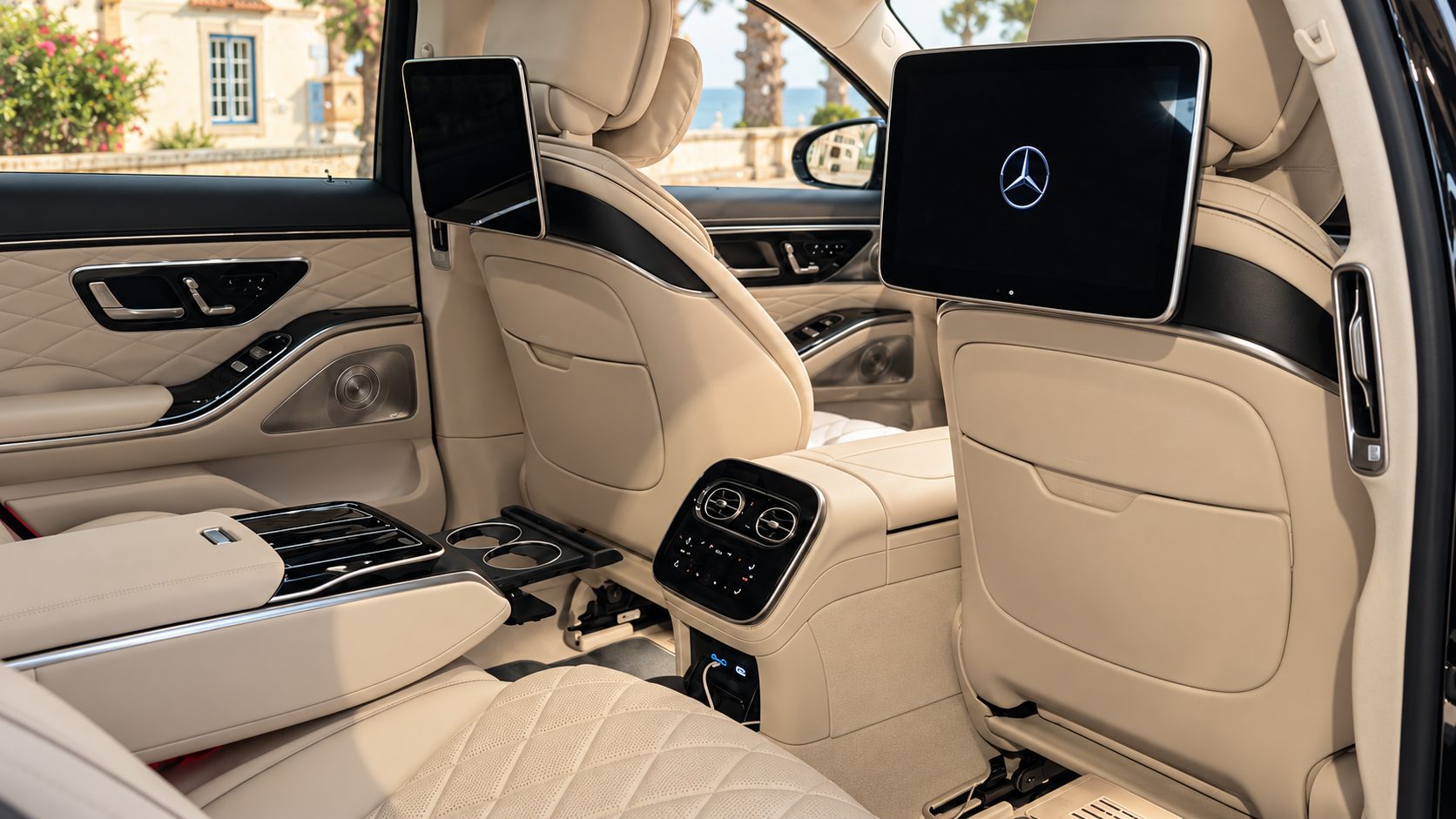 Chauffeur service Lisbon: rear cabin of a Mercedes-Benz S-Class with beige leather seats and screens in the seat backs, a villa and the sea visible through the window
