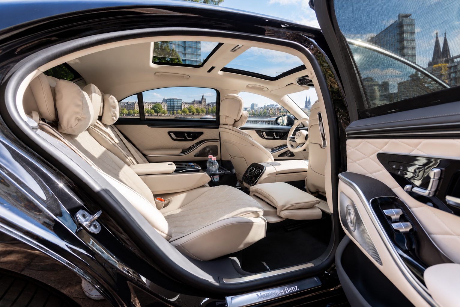 Chauffeurservice Köln: geöffnete Fondtür einer Mercedes-Benz S-Klasse mit hellen Ledersitzen, Fußstütze und Panoramadach, durch die Fenster der Rhein und der Kölner Dom