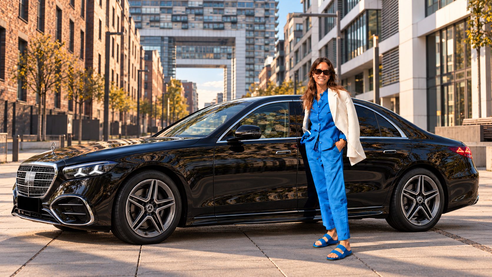 Frau in blauem Anzug mit weißem Blazer neben einer schwarzen Mercedes-Benz S-Klasse im Rheinauhafen in Köln, dahinter ein Kranhaus