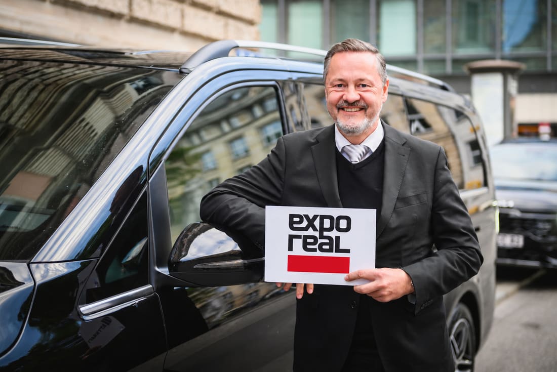AYDEN chauffeur in a dark suit leaning against a black Mercedes-Benz V-Class and holding an Expo Real pick-up sign