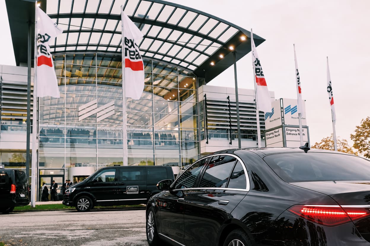 Black Mercedes-Benz S-Class outside the North Entrance of Messe München, with Expo Real flags on the flagpoles