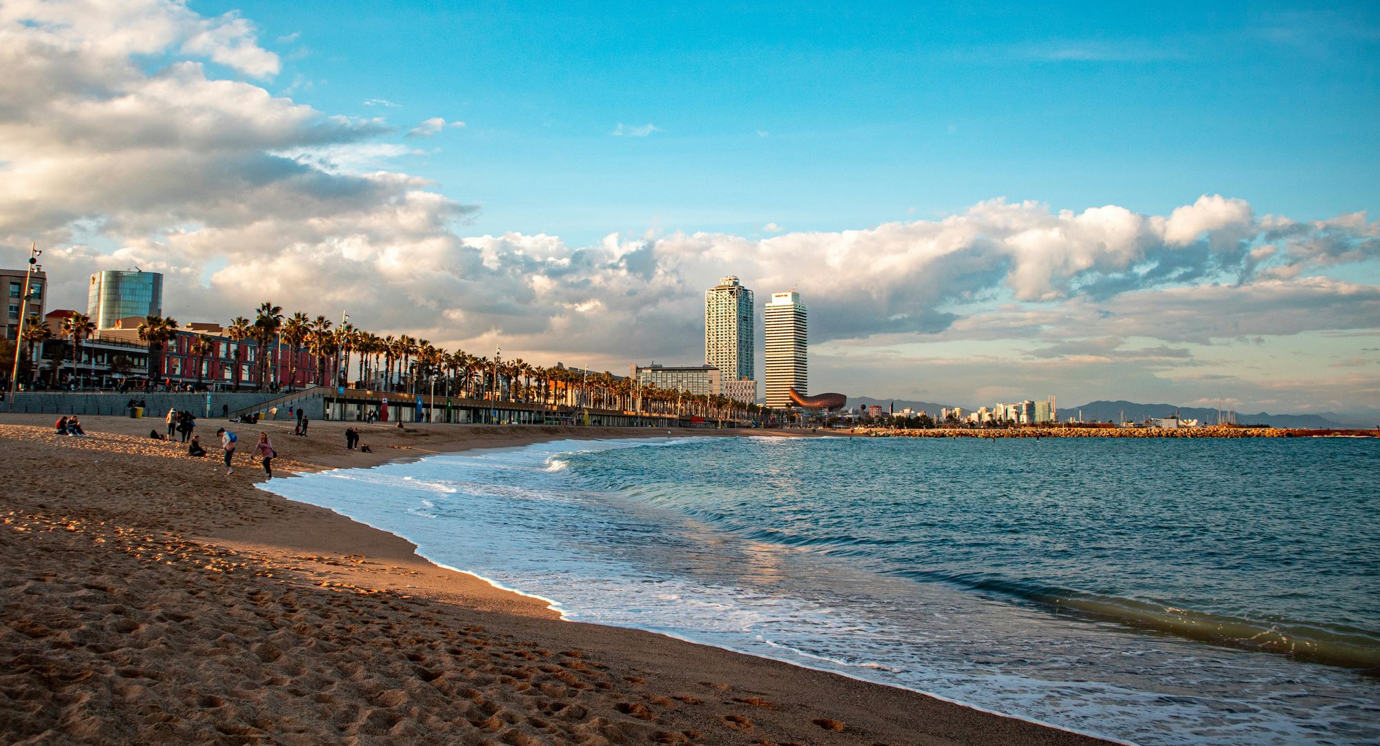 Strand Barceloneta in Barcelona mit Wellen, Palmen an der Promenade und den Türmen von Hotel Arts und Torre Mapfre am Port Olímpic unter Wolken