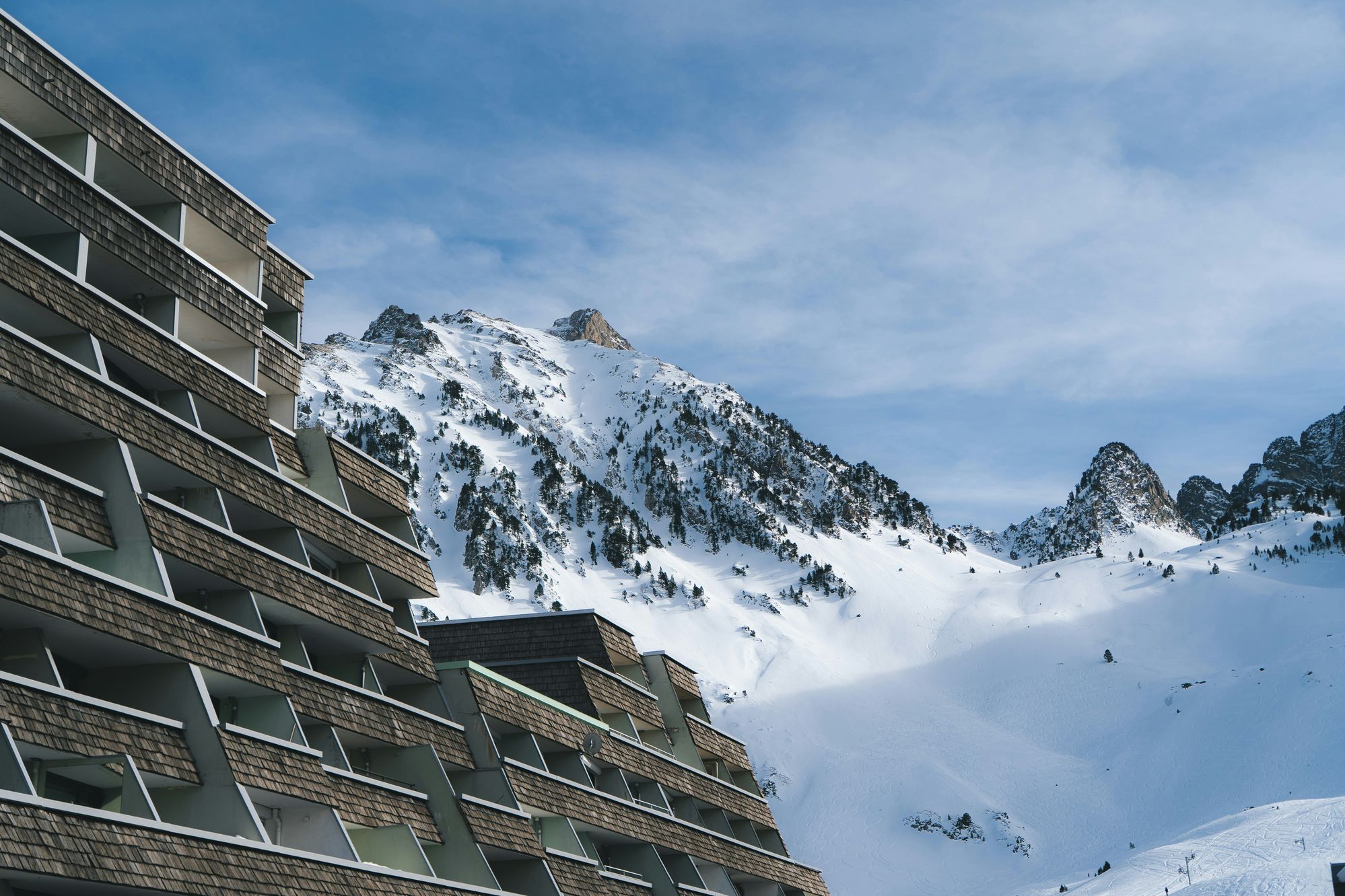 Hotelfassade mit Holzschindeln vor verschneiten Berggipfeln und Skipisten