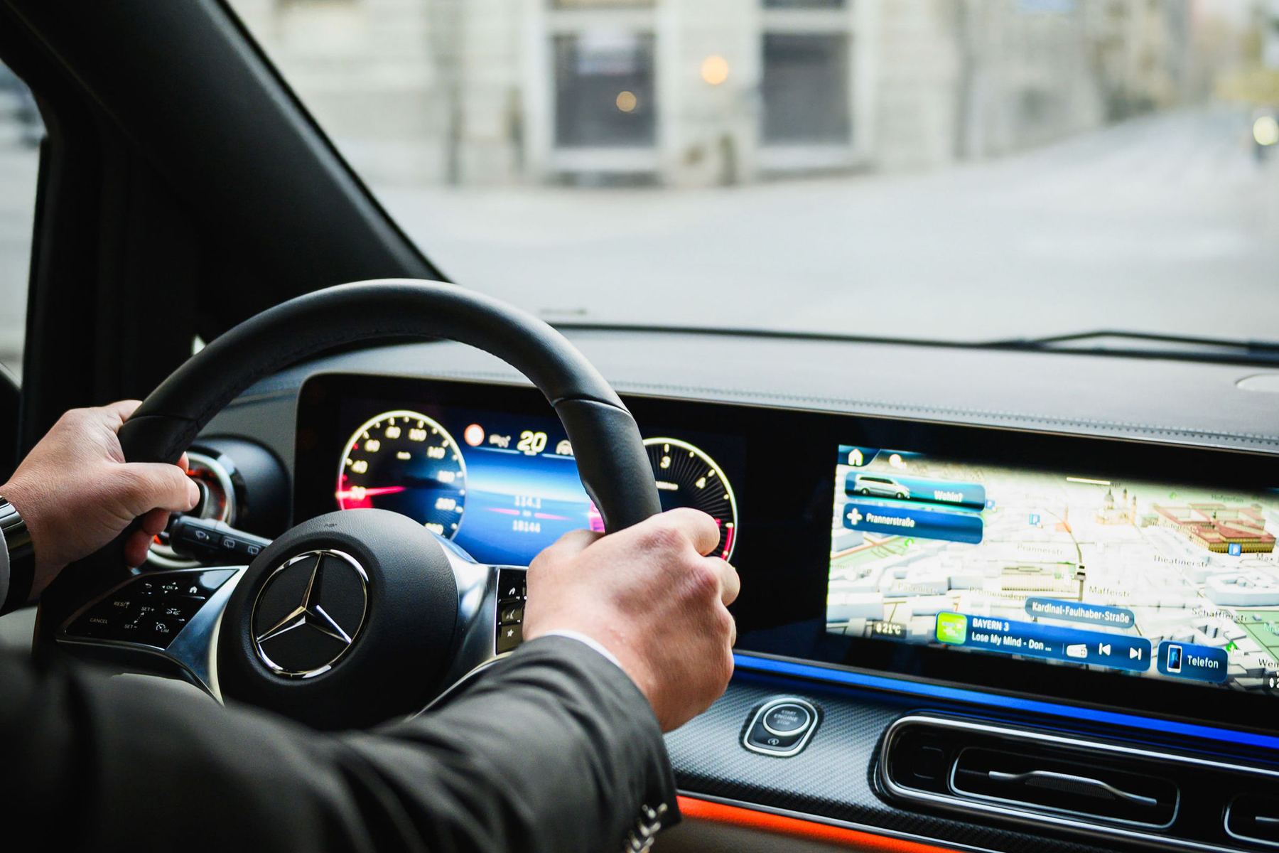 Chauffeur im Anzug am Lenkrad eines Mercedes-Benz, auf dem Bildschirm die Navigation durch die Münchner Innenstadt