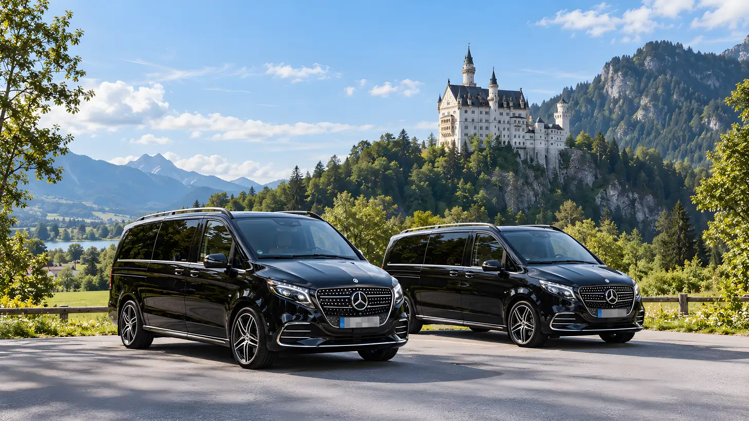 Zwei schwarze Mercedes-Benz V-Klassen auf einem Parkplatz unterhalb von Schloss Neuschwanstein, dahinter bewaldete Berge