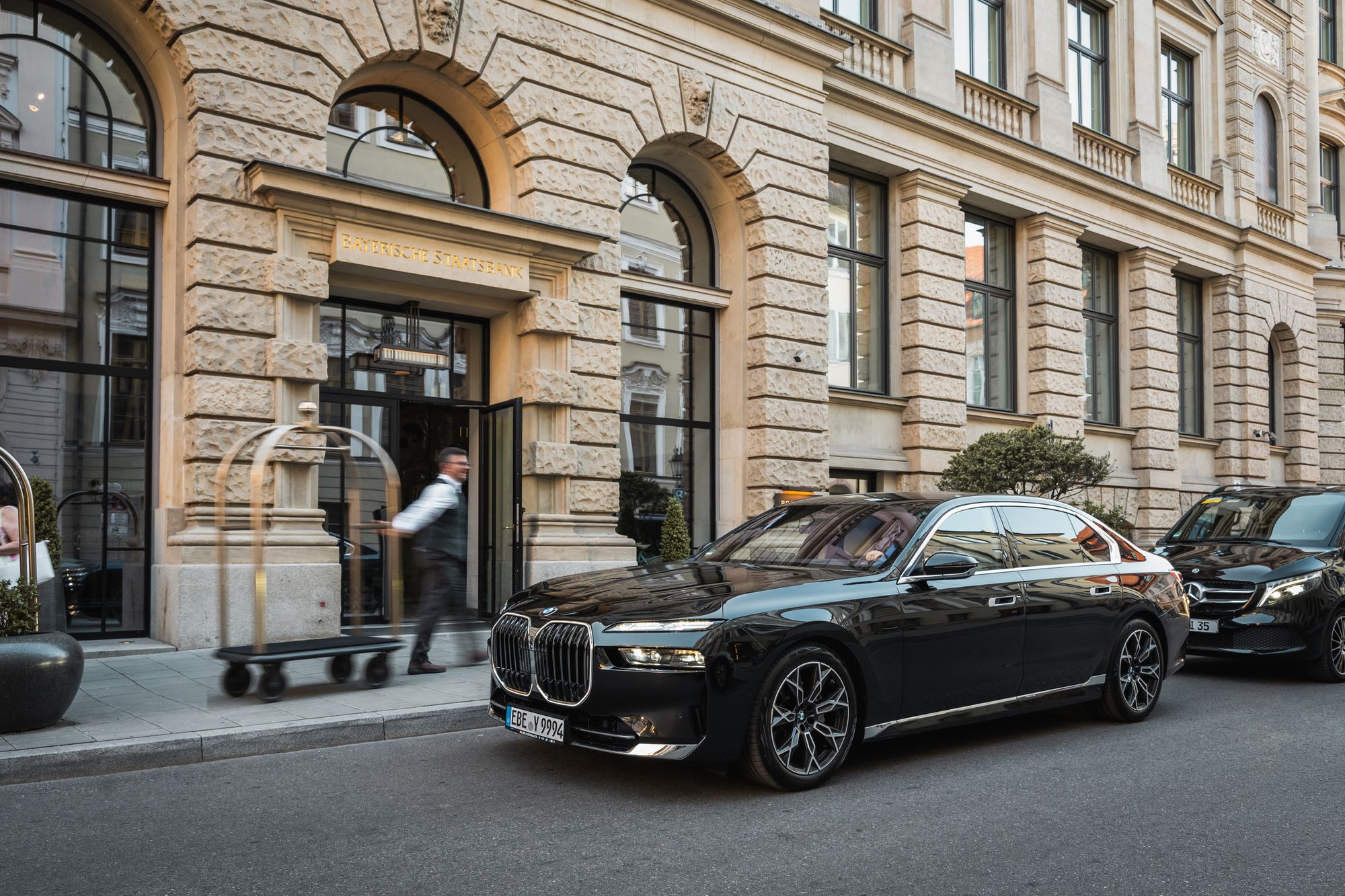 Black AYDEN BMW 7 Series outside a hotel entrance in Munich's old town, with a Mercedes V-Class behind it
